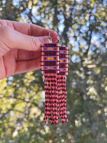 A pair of handmade beaded fringe earrings held by a hand against a blurred outdoor background of green trees and sunlight. The earrings feature a geometric pattern in purple, yellow, and white beads, with long fringe strands in dark red and pink.
