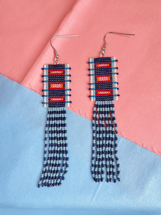 A pair of handmade beaded dangle earrings with silver fishhook wires, featuring geometric patterns in red, white, and dark blue, with long fringe strands. The earrings are displayed on a diagonally split pink and light blue fabric background.