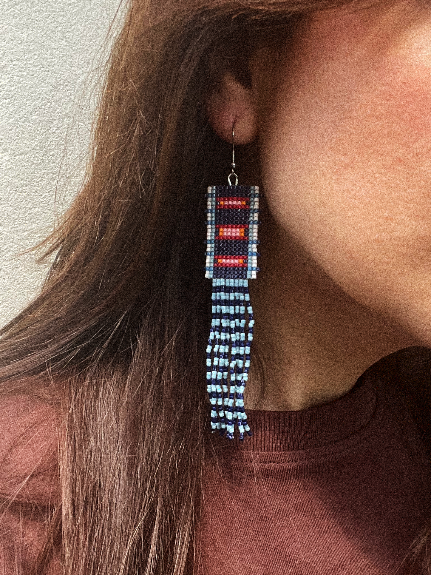 A close-up of a person wearing a long beaded dangle earring. The earring features a rectangular top section with red and white beadwork, connected to a long fringed drop made of light blue and white beads.