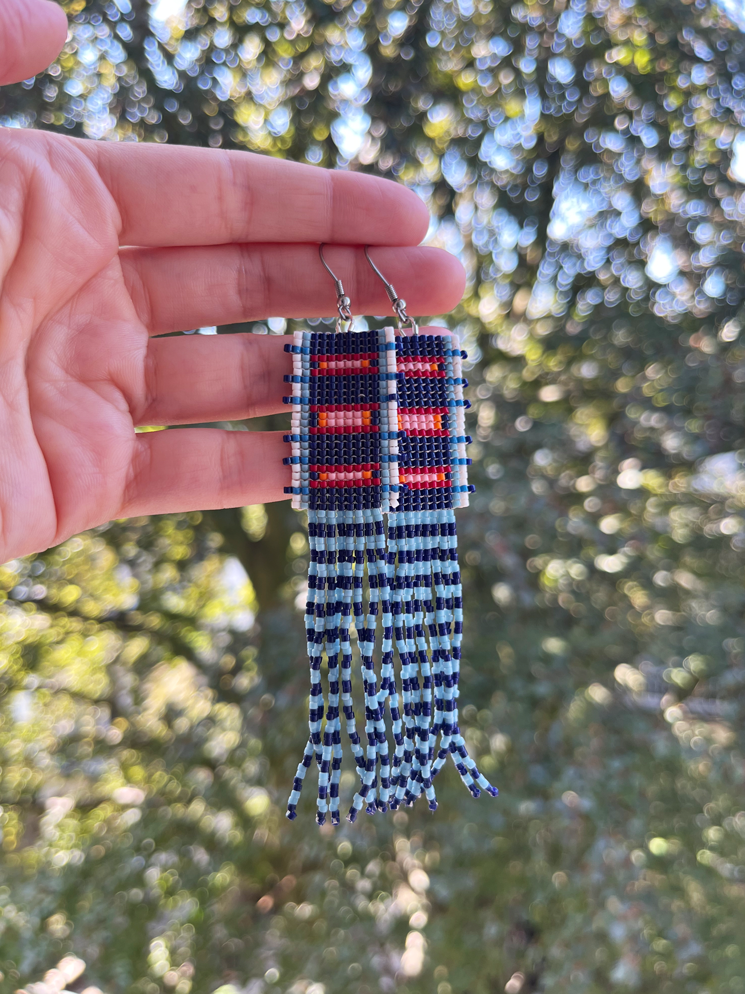 A pair of handmade beaded fringe earrings held up by a hand against a blurred outdoor background of green trees. The earrings feature a geometric pattern in navy blue, red, and white, with long fringe strands hanging below.