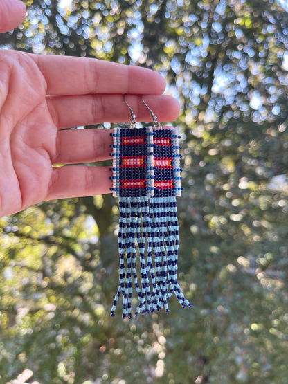 A pair of handmade beaded fringe earrings held up by a hand against a blurred outdoor background of green trees. The earrings feature a geometric pattern in navy blue, red, and white, with long fringe strands hanging below.