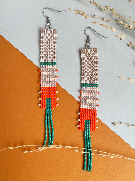 A pair of handmade beaded dangle earrings with fishhook wires, featuring geometric patterns in white, brown, green, orange, and red, with long green bead tassels. The earrings are displayed on a diagonally split gray and orange background with dried decorative grass.