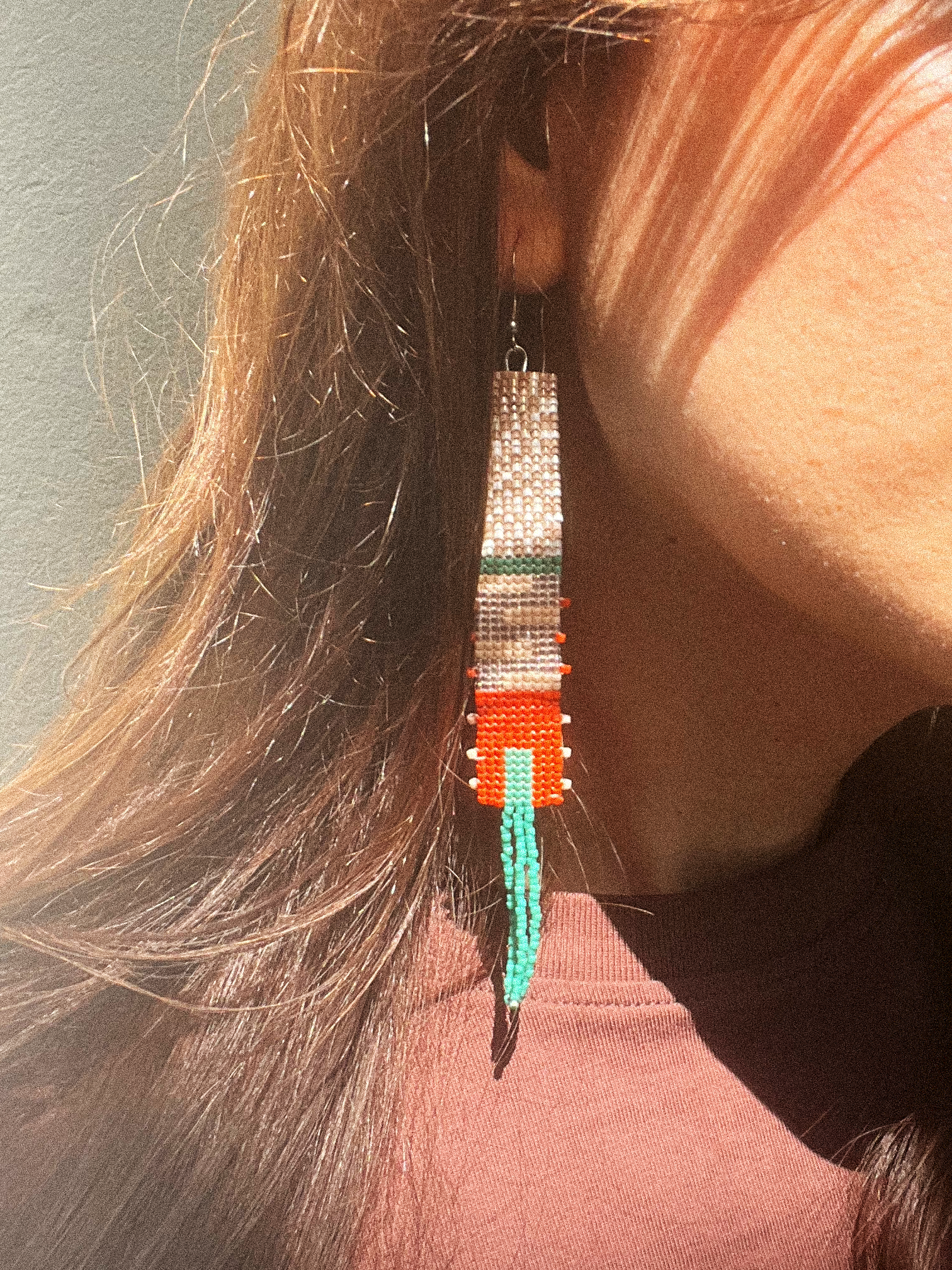 A close-up lifestyle shot of a person wearing a long, hand-beaded dangle earring. The earring features a white and green patterned top section, a bright orange middle section, and a turquoise blue beaded tassel at the bottom.