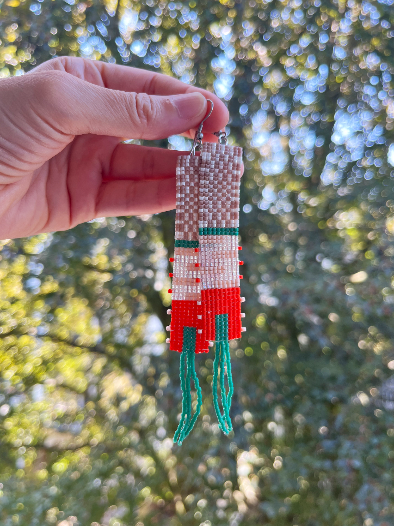 A hand holds a long, hand-beaded earring outdoors against a blurred background of green trees and sunlight. The earring features a geometric pattern in white, red, and green seed beads with a silver fishhook ear wire.