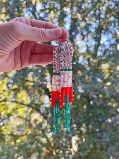 A hand holds a long, hand-beaded earring outdoors against a blurred background of green trees and sunlight. The earring features a geometric pattern in white, red, and green seed beads with a silver fishhook ear wire.