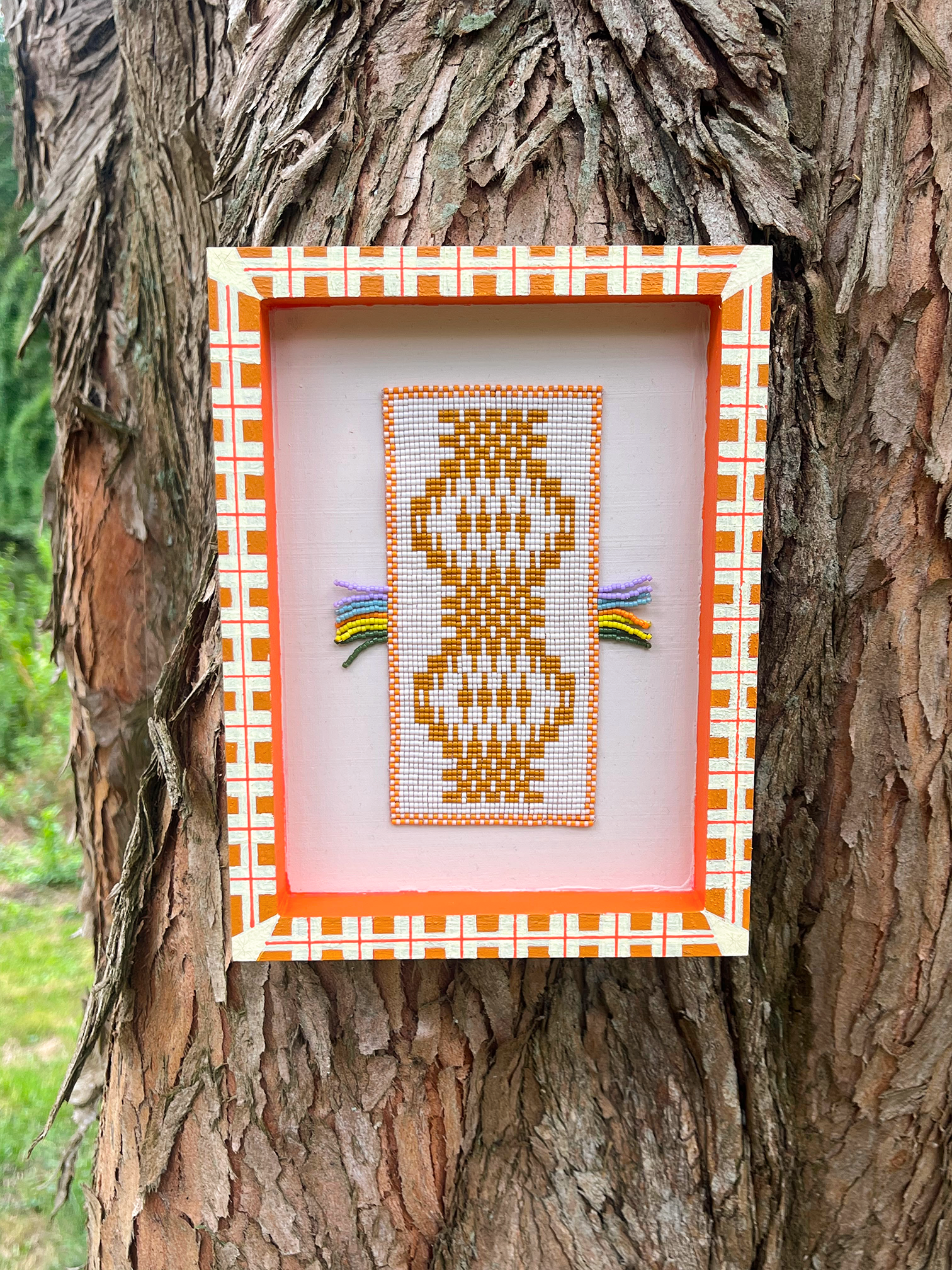 Framed beaded wall panel with mustard folk figure motif and rainbow fringe, resting on tree bark outdoors