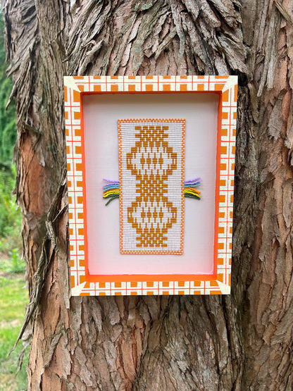 Framed beaded wall panel with mustard folk figure motif and rainbow fringe, resting on tree bark outdoors
