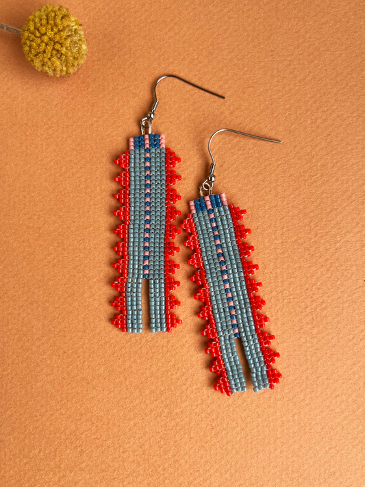 A pair of handmade beaded dangle earrings with a geometric pattern in red, light blue, and pink, displayed on a textured orange background with a small yellow flower prop.