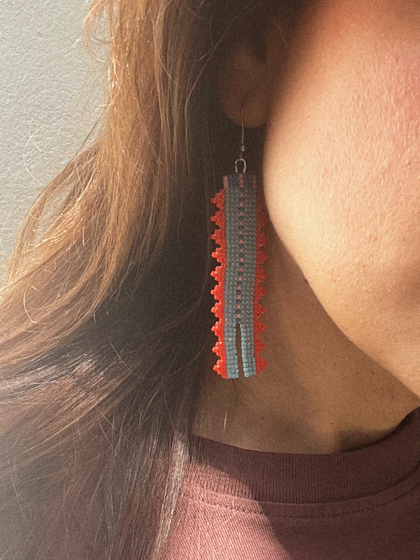 A close-up of a person wearing a long, rectangular beaded dangle earring in red, blue, and gray, with long brown hair and a mauve top visible.