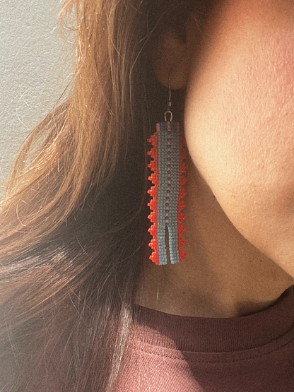 A close-up of a person wearing a long, rectangular beaded dangle earring in red, blue, and gray, with long brown hair and a mauve top visible.