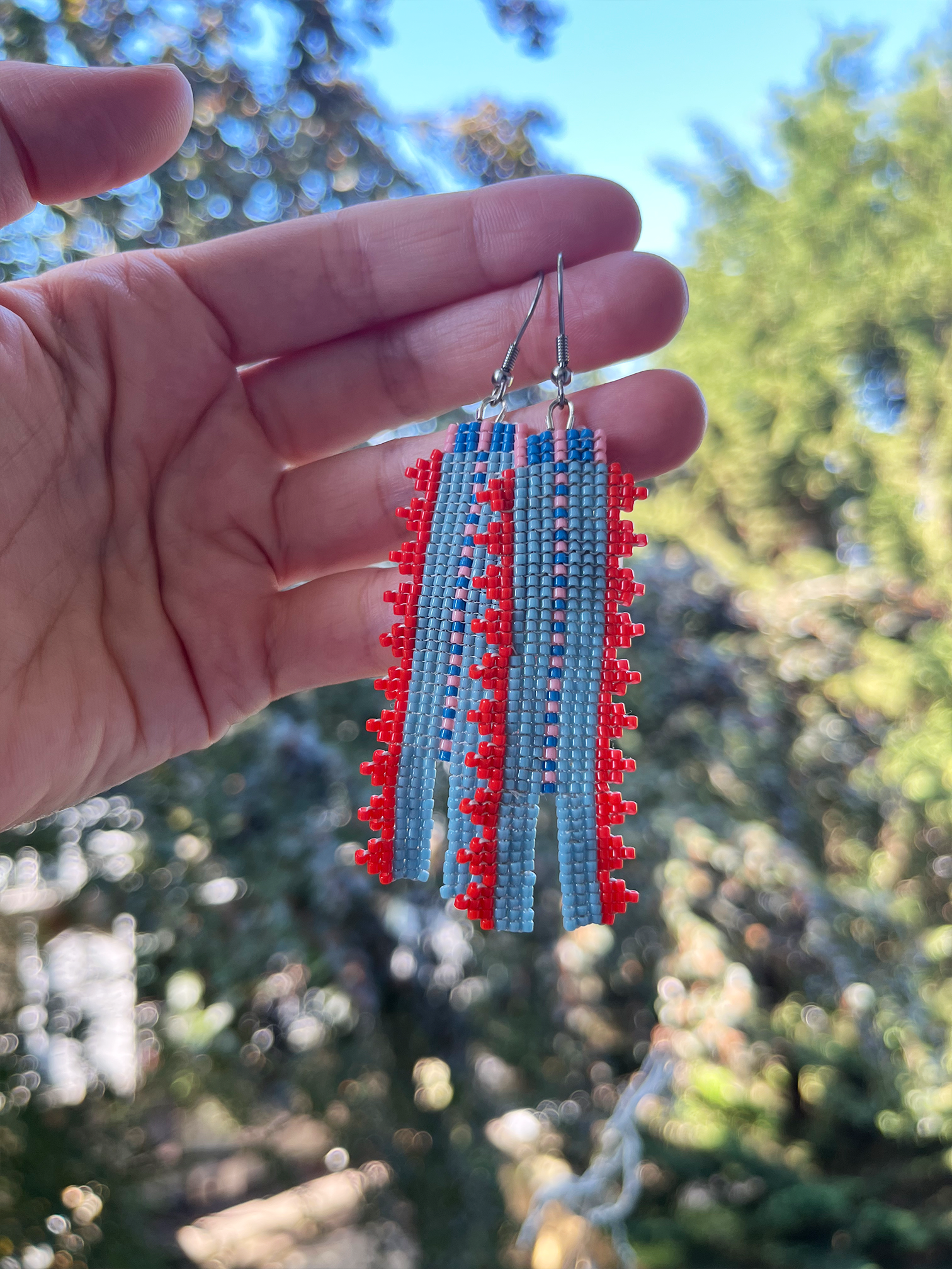 A pair of handmade beaded dangle earrings held in a hand against a blurred outdoor background of trees and blue sky. The earrings feature light blue and red beads in a geometric pattern with silver-toned hooks.