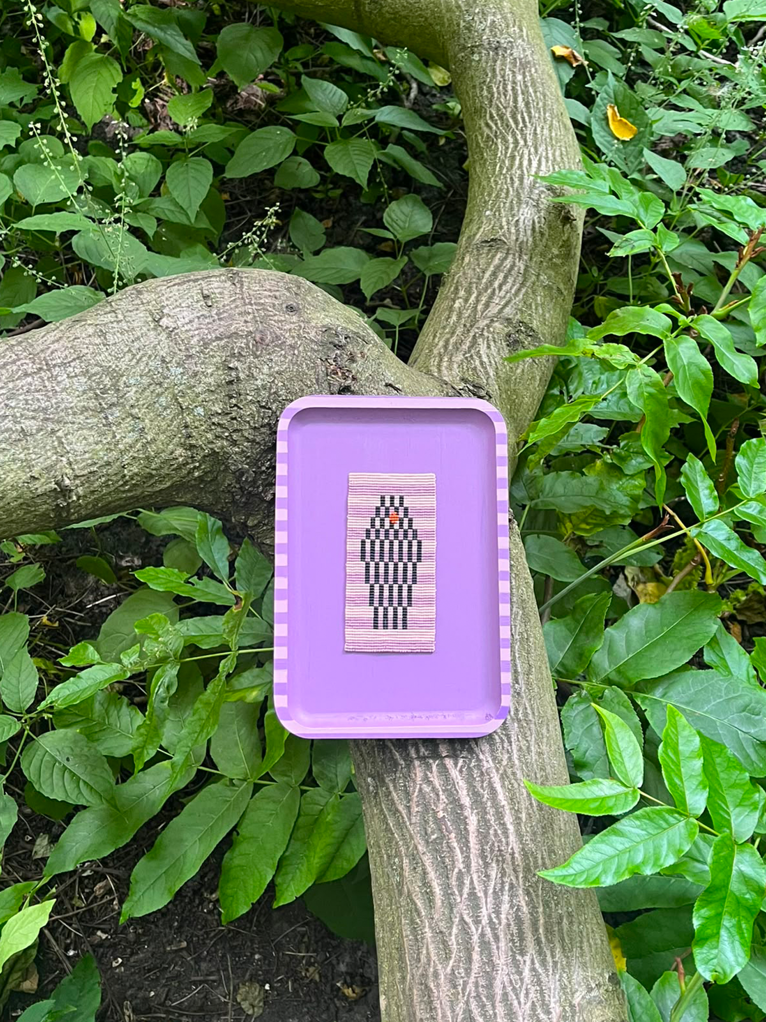 A lavender purple rectangular tray with a striped border sits on a tree branch outdoors, surrounded by green leaves. The tray holds a small square beaded piece featuring a  geometric design with a small orange accent.
