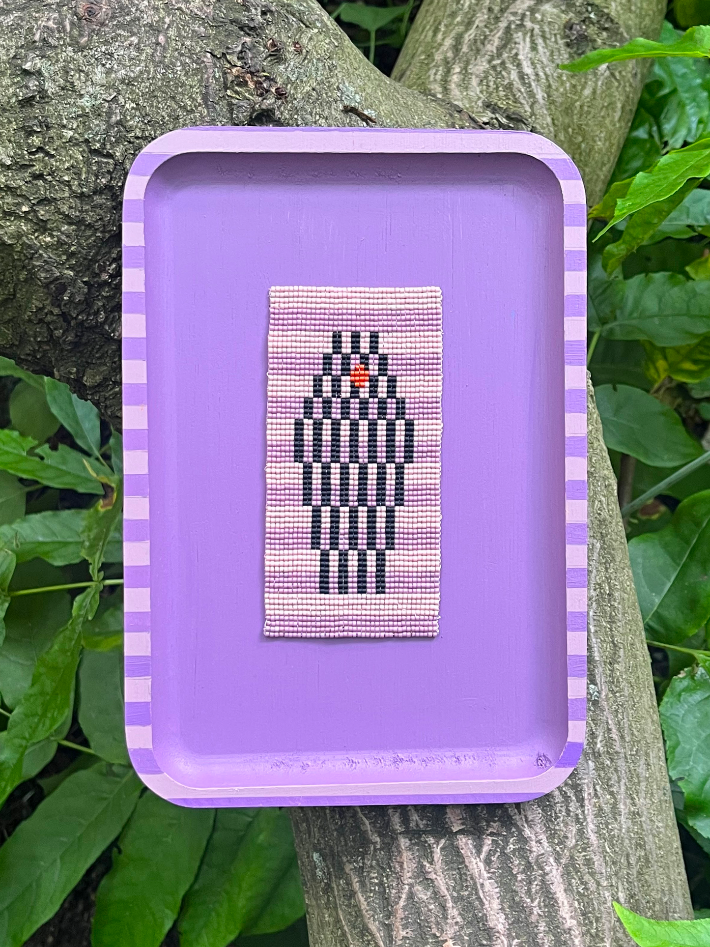 The Watcher beaded panel with black diamond motif on pink, framed in lavender, resting against tree bark