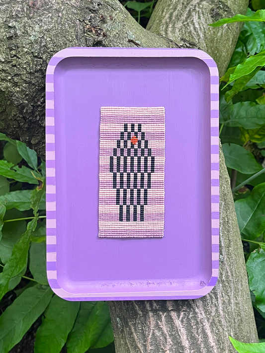 The Watcher beaded panel with black diamond motif on pink, framed in lavender, resting against tree bark