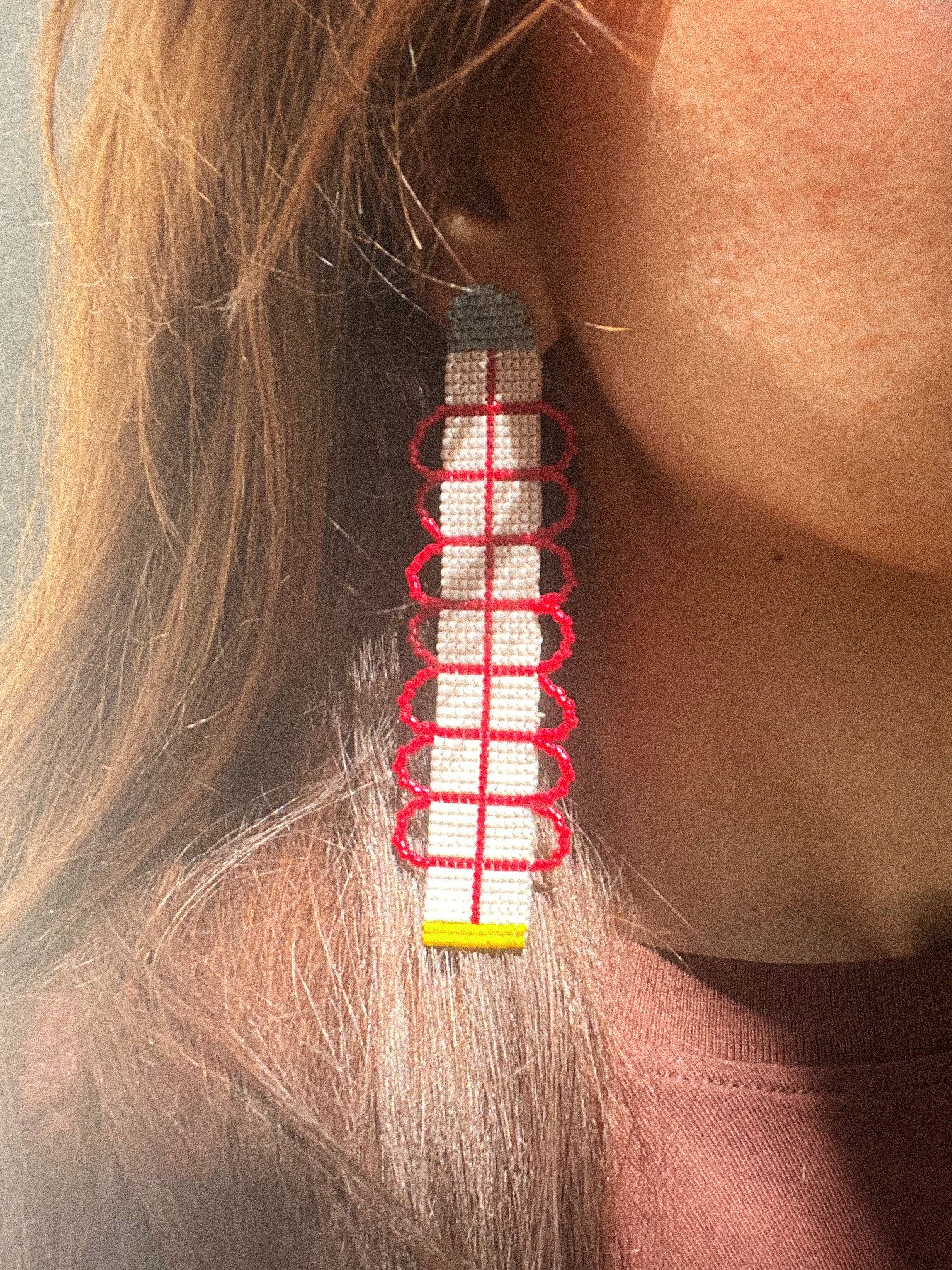 A close-up of a person wearing a long, geometric beaded earring. The earring features white woven beads, red geometric wire-like accents, a gray top section, and a small yellow bottom accent.