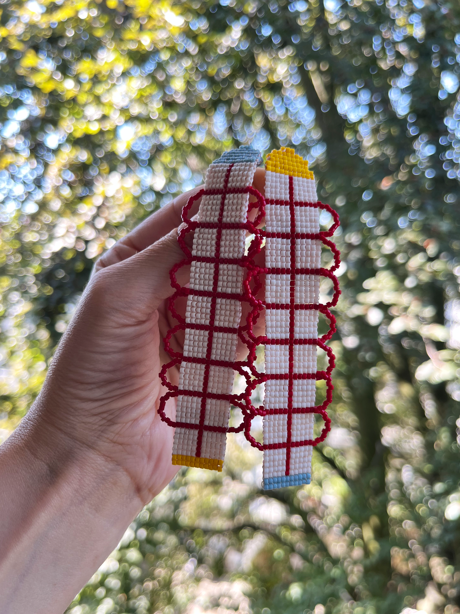 A hand holds two handmade woven belts outdoors against a blurred background of green trees and sunlight. The belts are primarily white and red with small yellow and blue accents.