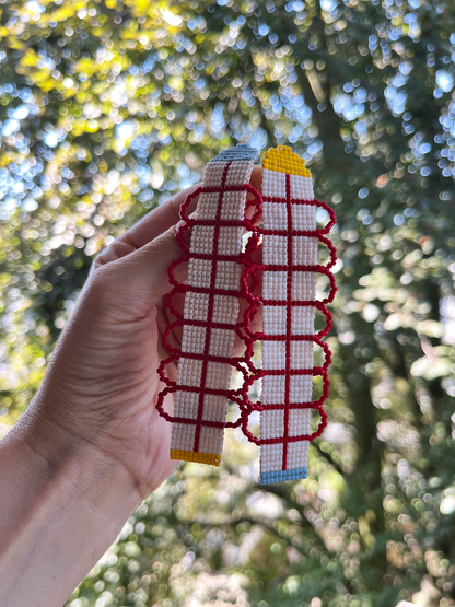 A hand holds two handmade woven belts outdoors against a blurred background of green trees and sunlight. The belts are primarily white and red with small yellow and blue accents.