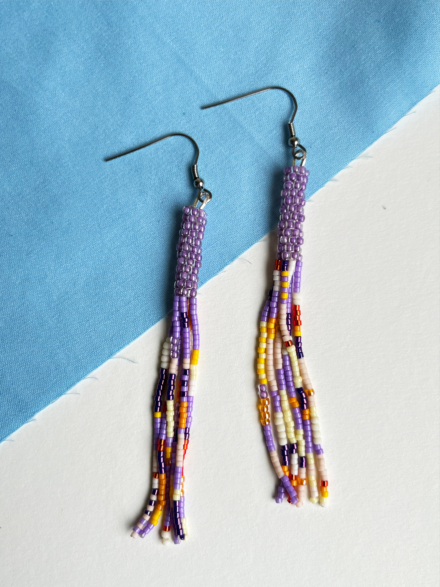 A pair of handmade beaded tassel earrings with silver fishhook wires, featuring purple, white, yellow, and dark purple seed beads, displayed on a diagonally split blue and white surface.