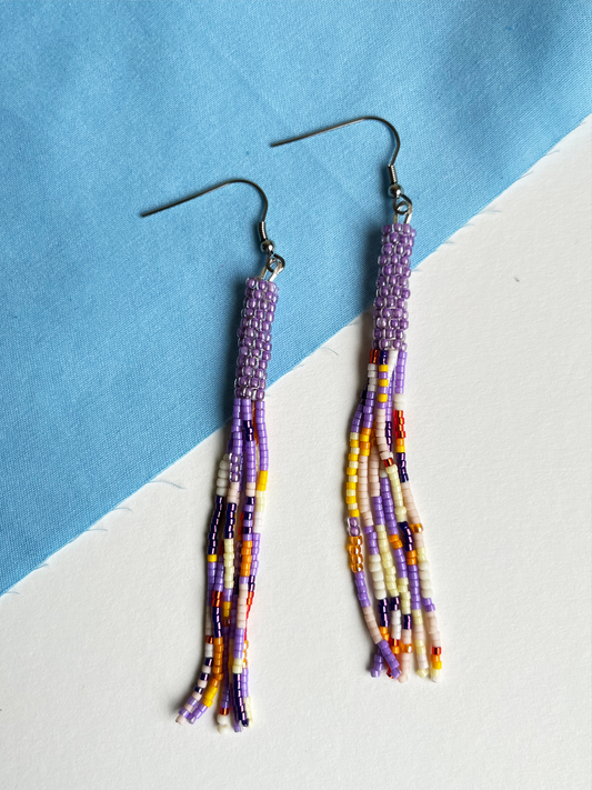 A pair of handmade beaded tassel earrings with silver fishhook wires, featuring purple, white, yellow, and dark purple seed beads, displayed on a diagonally split blue and white surface.