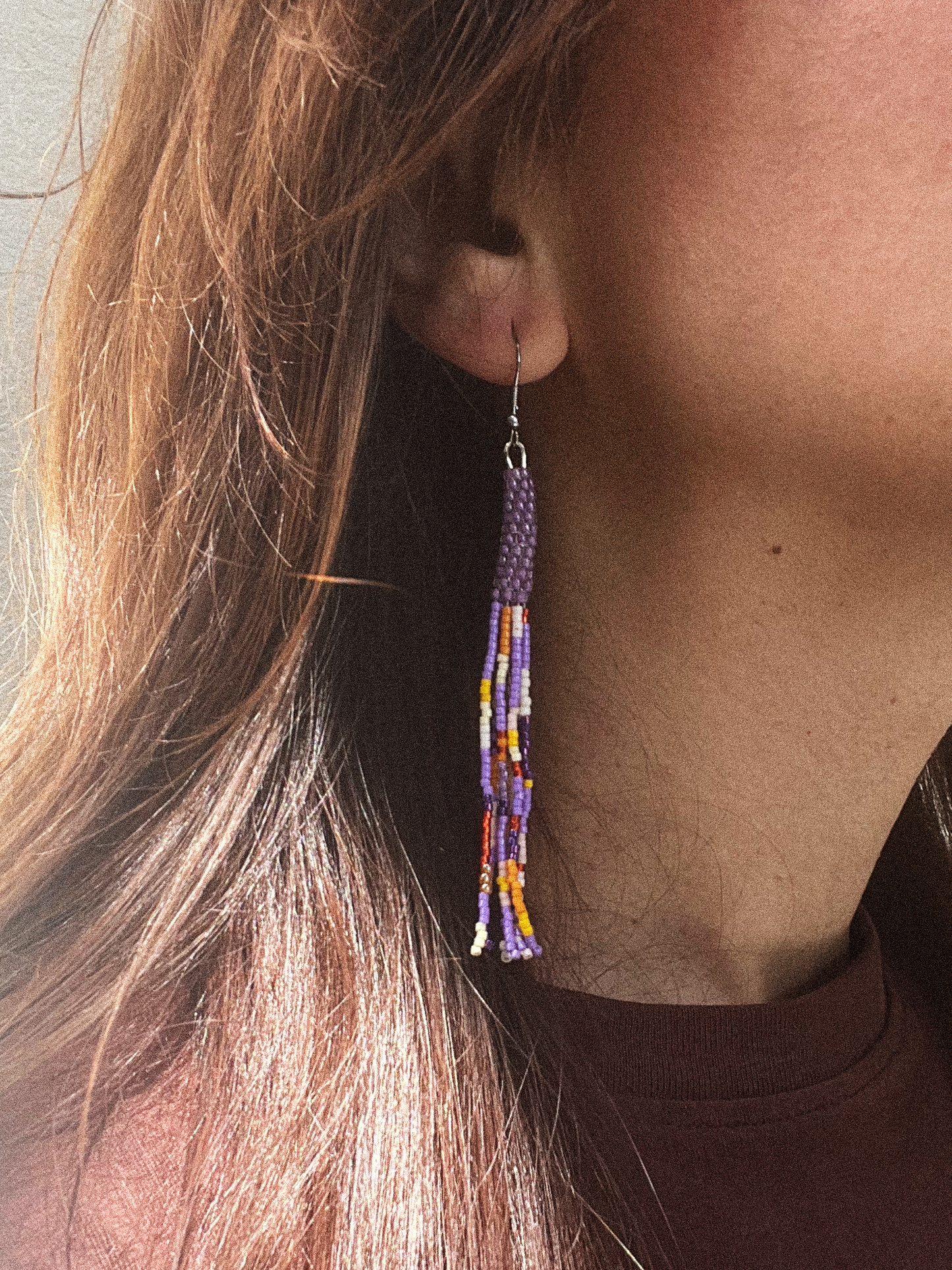A close-up of a person's ear wearing a long beaded dangle earring. The earring features a purple woven top section with dangling tassel-like strands in purple, white, orange, and yellow.