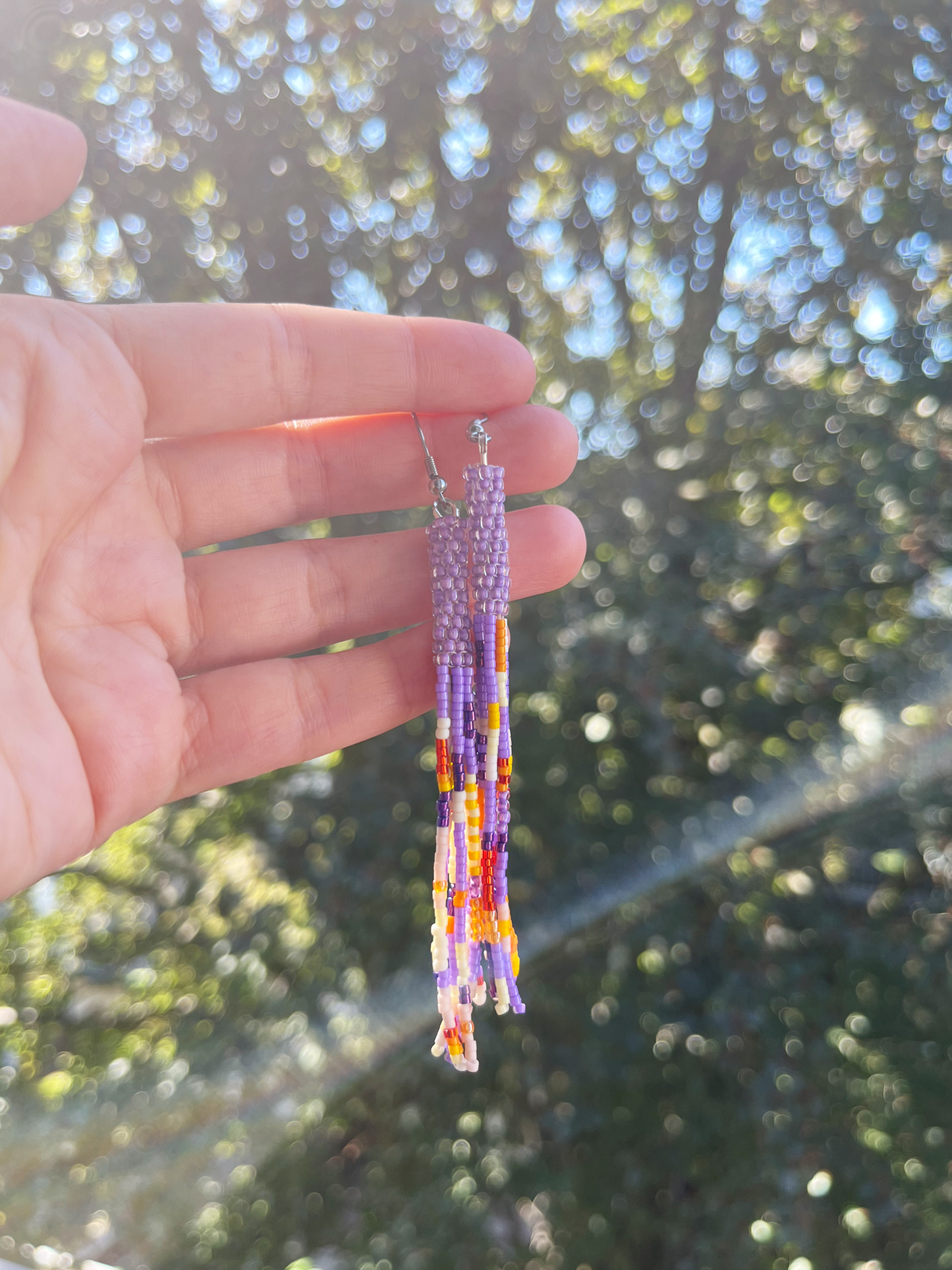 A hand holding a pair of long beaded dangle earrings outdoors, with a blurred background of green foliage and sunlight.