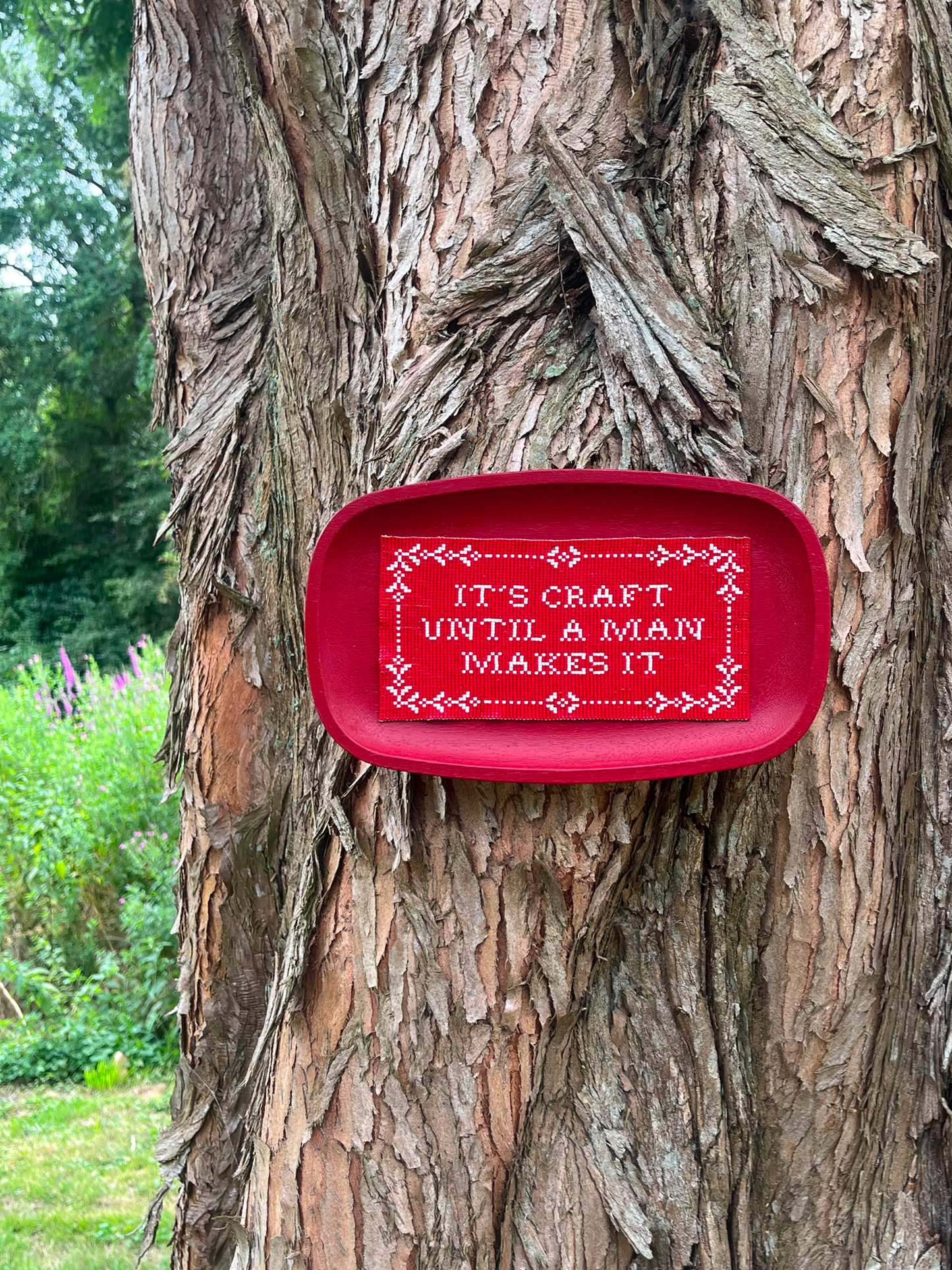 Red beaded sampler tray reading "It's craft until a man makes it" hanging on tree bark outdoors